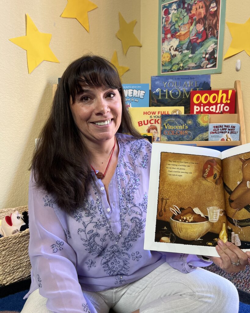 Leslie Johnson, Mountain Resource Center Preschool Assistant Director. A white woman with long, dark hair and a purple top sits on the floor and shows a book as if she is reading to children.