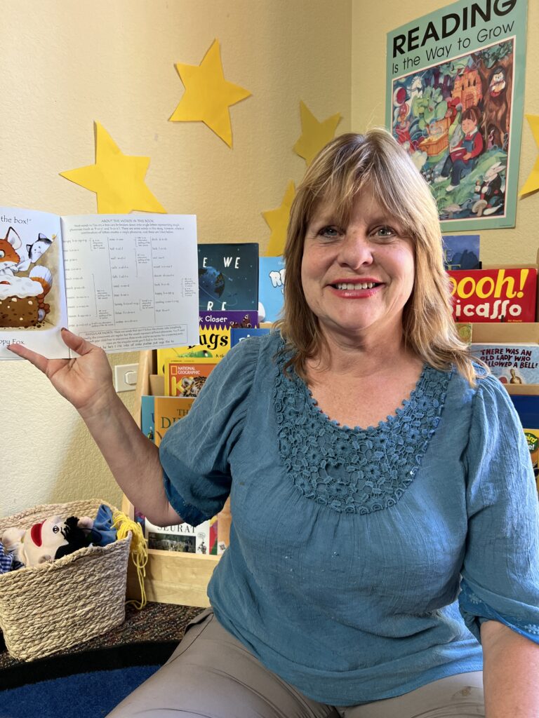 Carla Keitlen, Preschool Director. A blond-haired white woman wearing a blue top, holding up a book as if reading to children.